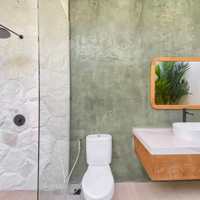 Modern bathroom with a wall-mounted rainfall shower, white stone wall, and green textured wall; wooden-framed mirror reflects plants.