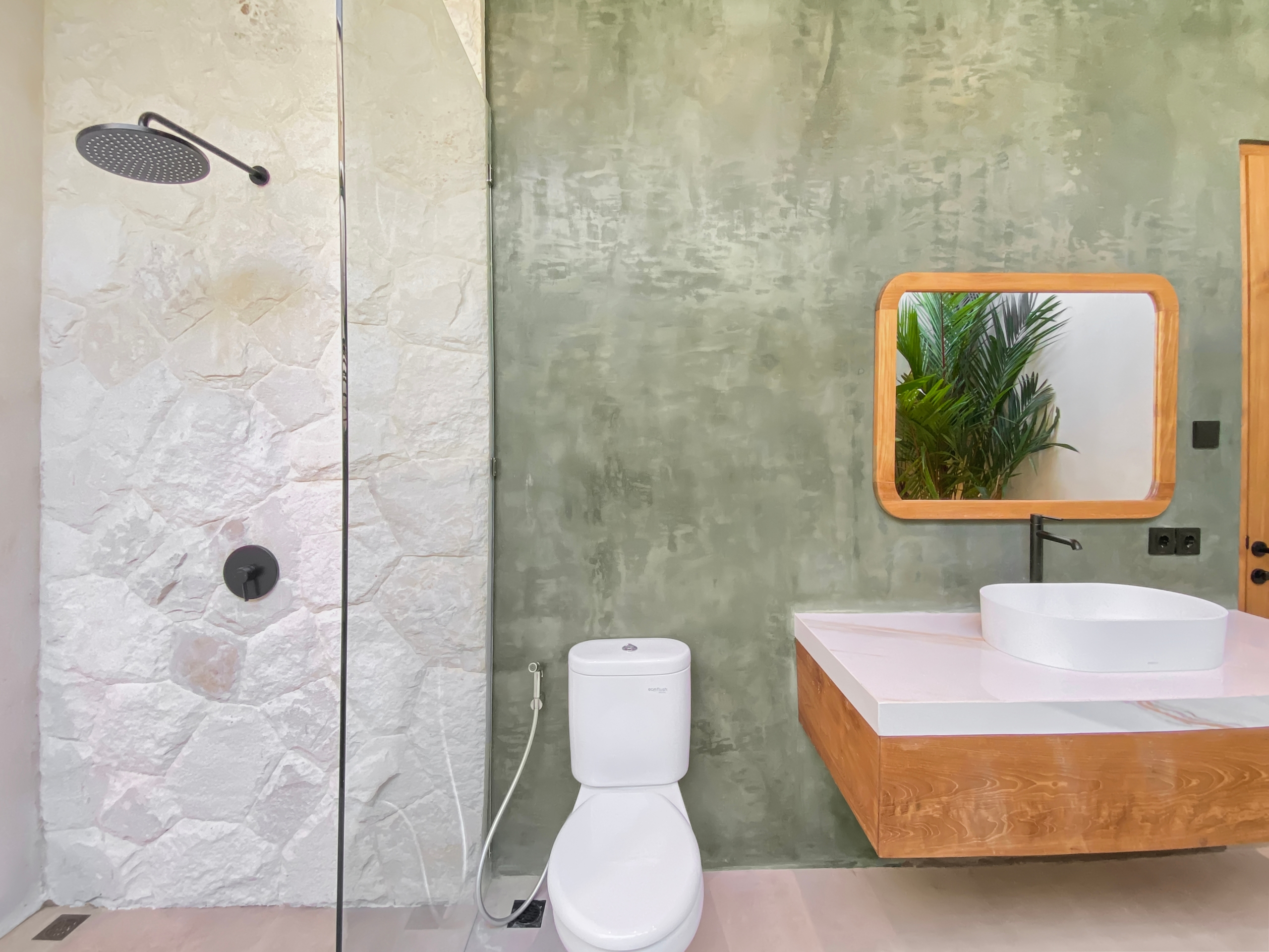 Modern bathroom with a wall-mounted rainfall shower, white stone wall, and green textured wall; wooden-framed mirror reflects plants.