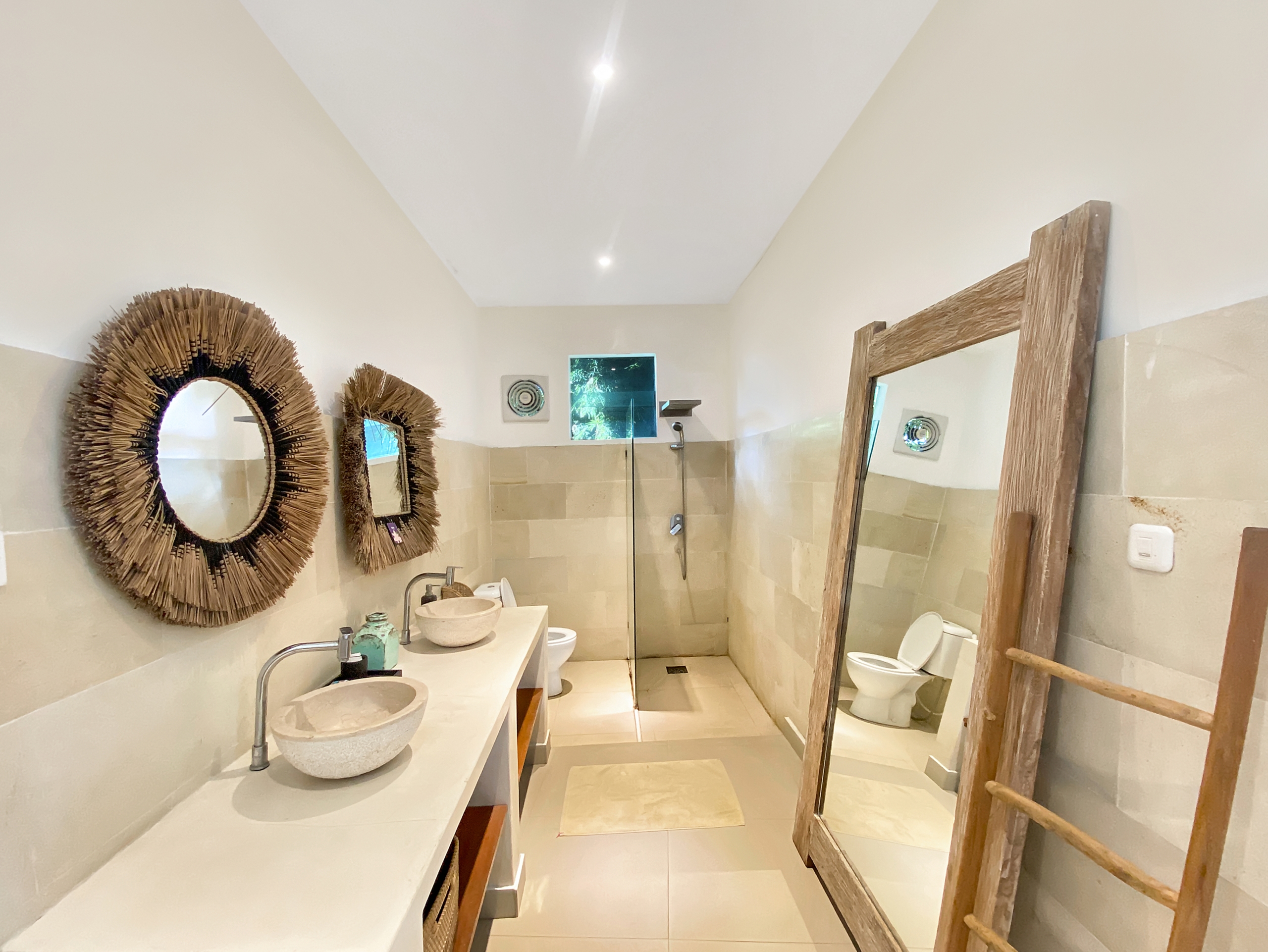 Bathroom with two stone sinks on a long counter, woven circular mirrors, and a wooden ladder mirror setup.