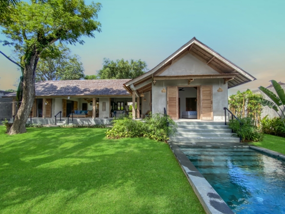 Front view of a single-story house with wooden shutters, surrounded by a well-kept green lawn and tropical plants; a narrow reflecting pool runs along the right side of the scene.