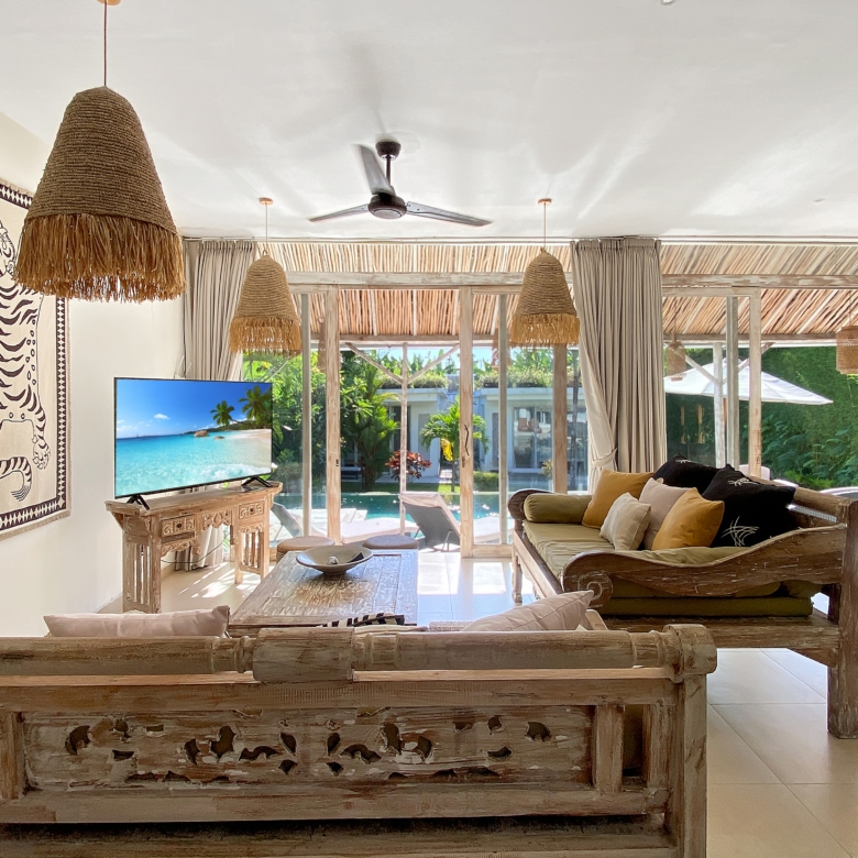 Bright tropical living room with rustic wooden furniture, large woven pendant lamps, and glass doors opening to a pool area outside.