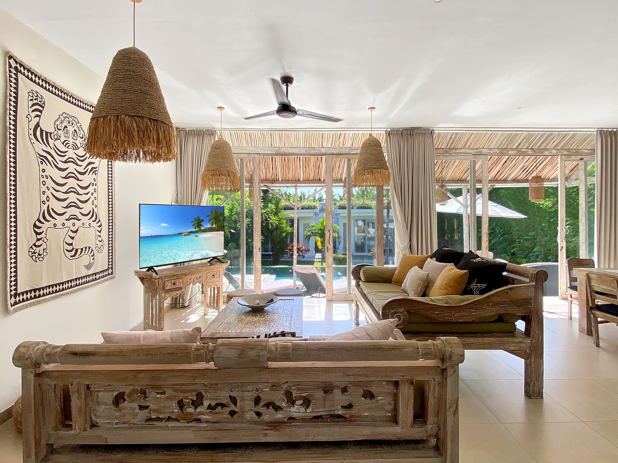 Bright tropical living room with rustic wooden furniture, large woven pendant lamps, and glass doors opening to a pool area outside.