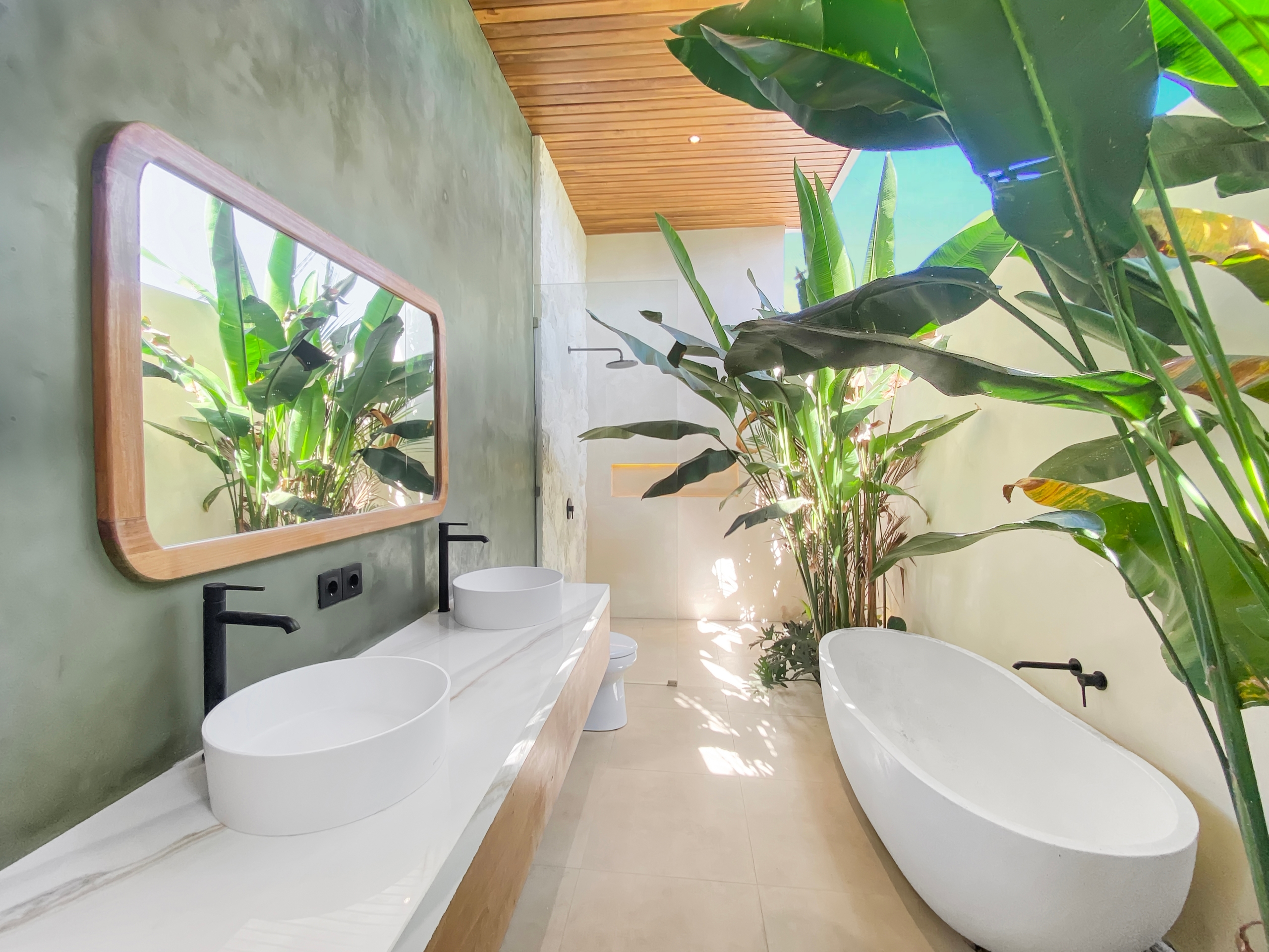 Modern bathroom with white freestanding tub, double vessel sinks, black fixtures, and large tropical plants near a green wall.