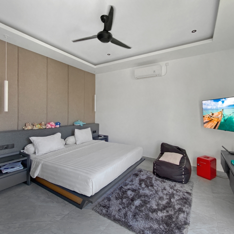 Modern hotel-style bedroom with a large bed, beige padded wall panels, gray headboard, ceiling fan, and wall-mounted TV. The room has a gray shag rug and a small desk.