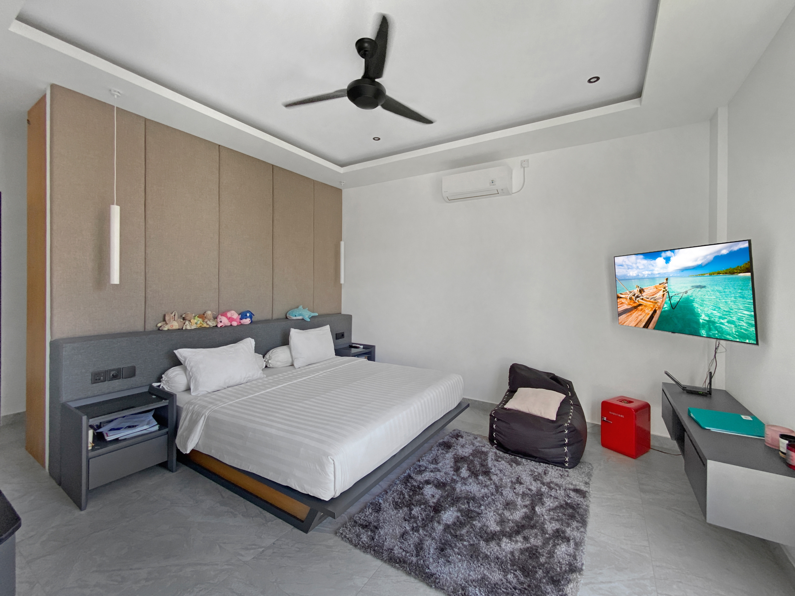 Modern hotel-style bedroom with a large bed, beige padded wall panels, gray headboard, ceiling fan, and wall-mounted TV. The room has a gray shag rug and a small desk.