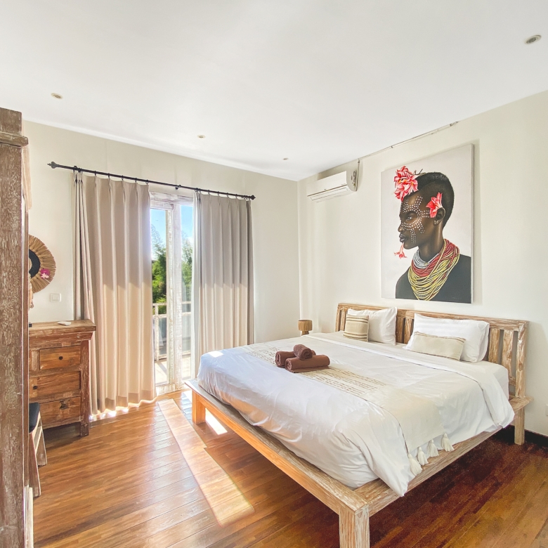 Rustic bedroom with a wooden bed and dresser, off-white walls, and a large portrait of a bead-adorned woman with pink flowers on the wall.