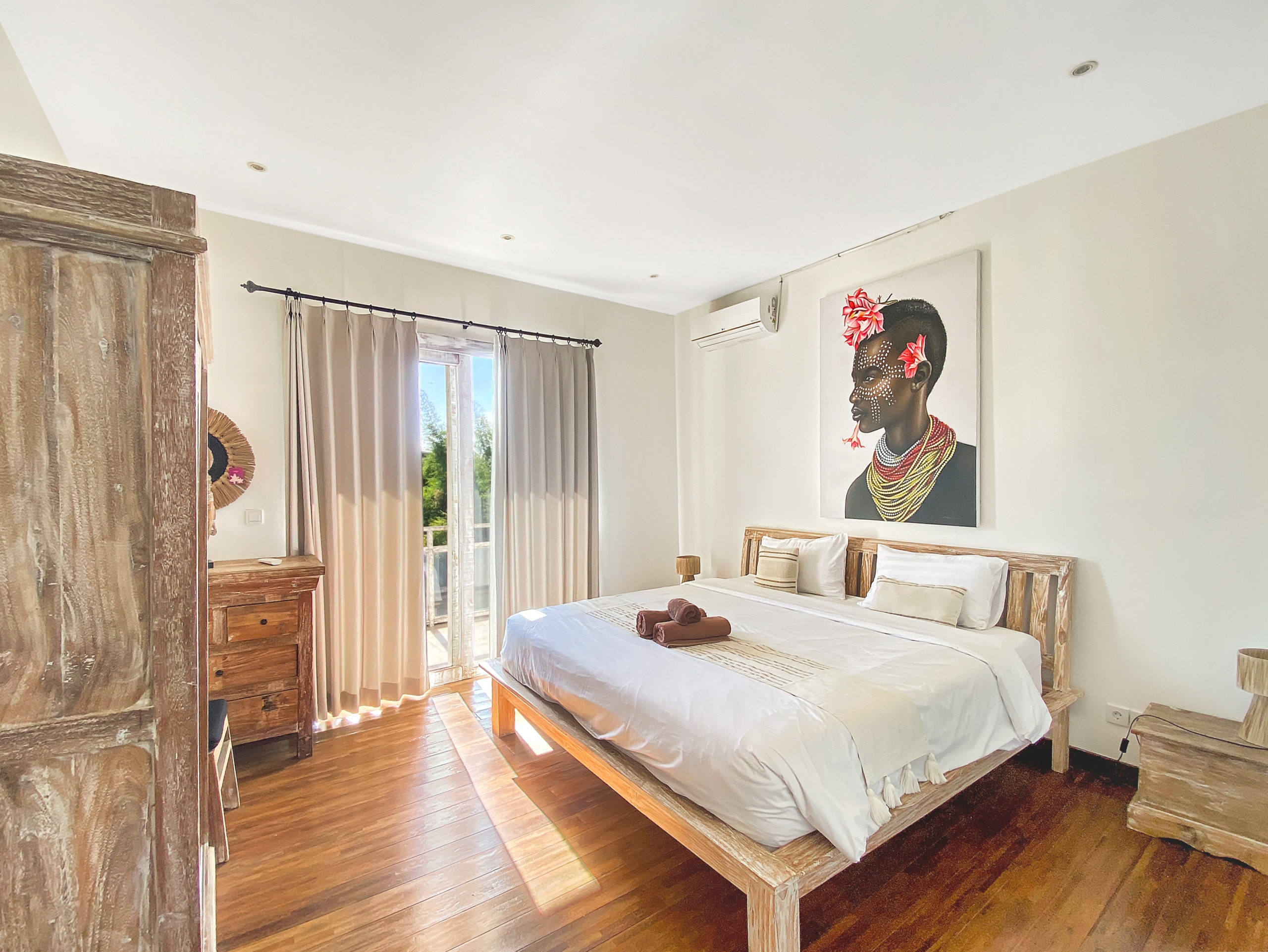 Rustic bedroom with a wooden bed and dresser, off-white walls, and a large portrait of a bead-adorned woman with pink flowers on the wall.
