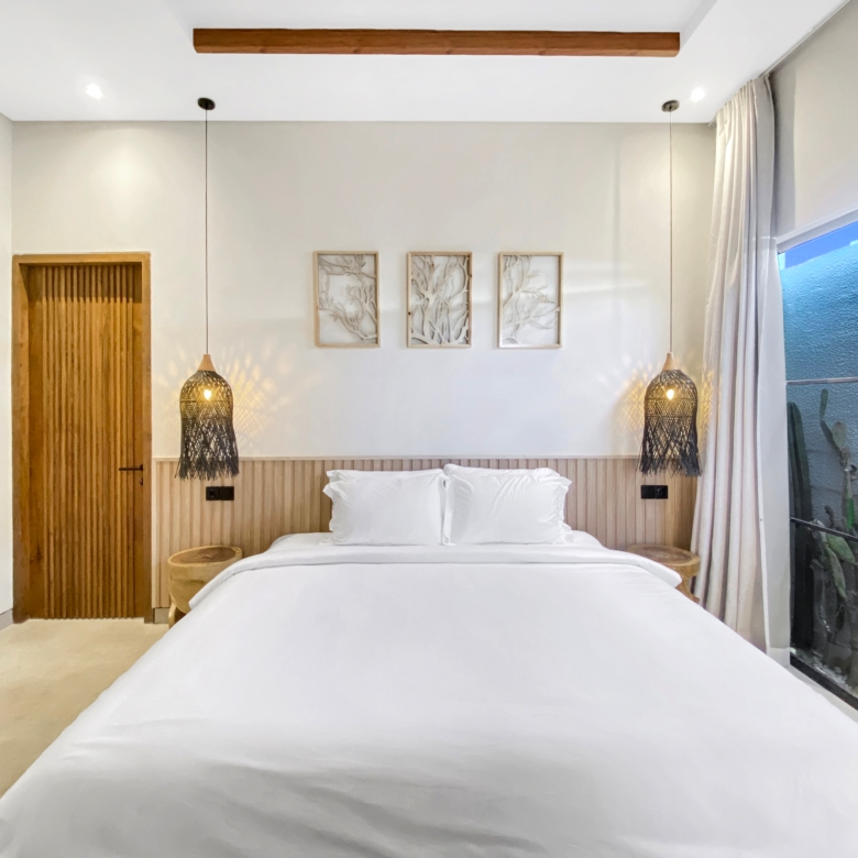 Bedroom with a single large bed in white linens, wooden headboard, and warm hanging lamps on each side; three framed botanical artworks above the headboard and a wooden door to the left, plus a glass wall with curtains on the right.