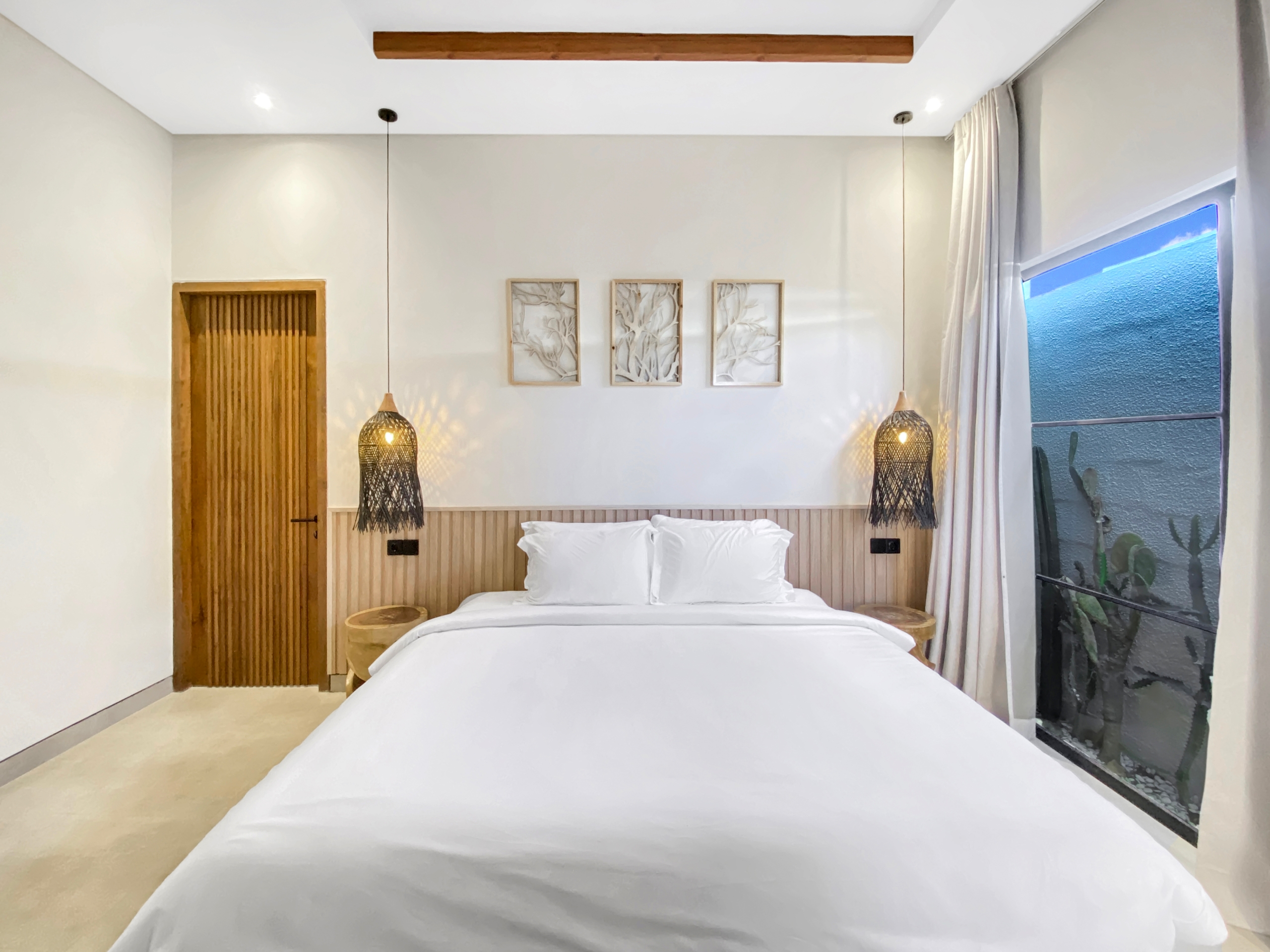 Bedroom with a single large bed in white linens, wooden headboard, and warm hanging lamps on each side; three framed botanical artworks above the headboard and a wooden door to the left, plus a glass wall with curtains on the right.