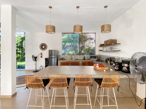 Open-plan kitchen with a long concrete island, five wicker bar stools, and three woven pendant lights overhead.