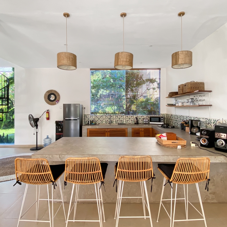 Open-plan kitchen with a long concrete island, five wicker bar stools, and three woven pendant lights overhead.