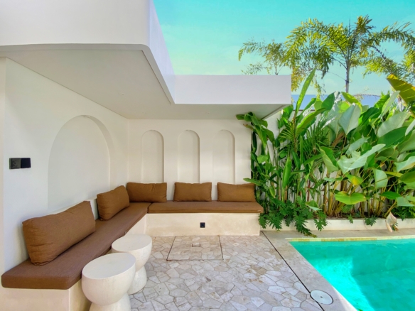 White minimalist poolside lounge with brown-cushioned seating and curved white walls next to tropical plants by a turquoise pool.