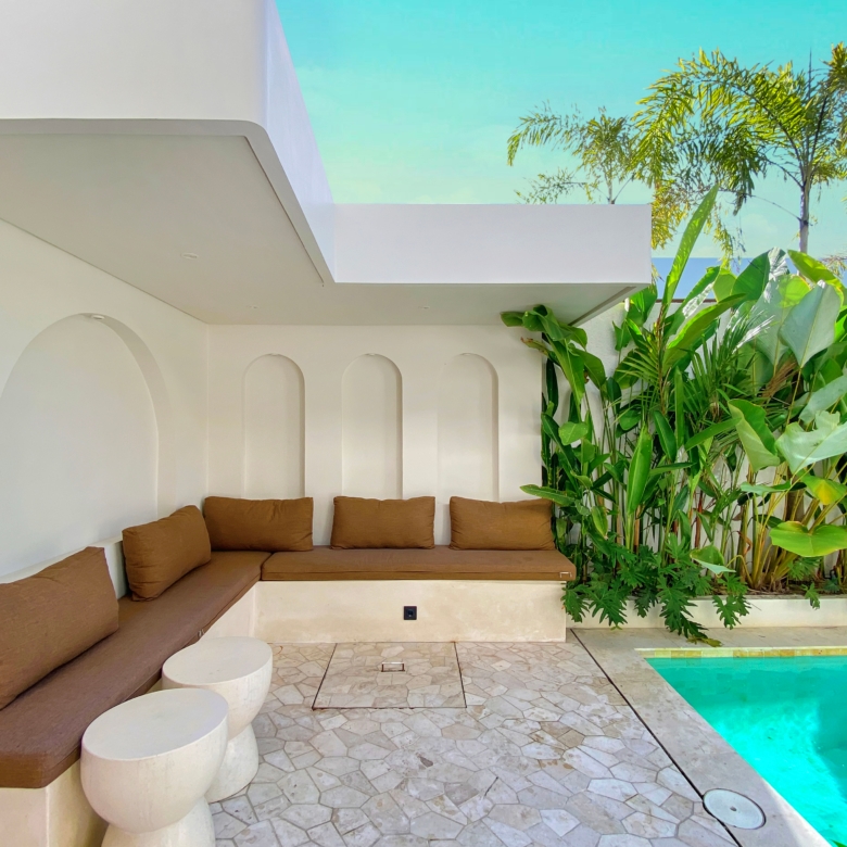 White minimalist poolside lounge with brown-cushioned seating and curved white walls next to tropical plants by a turquoise pool.