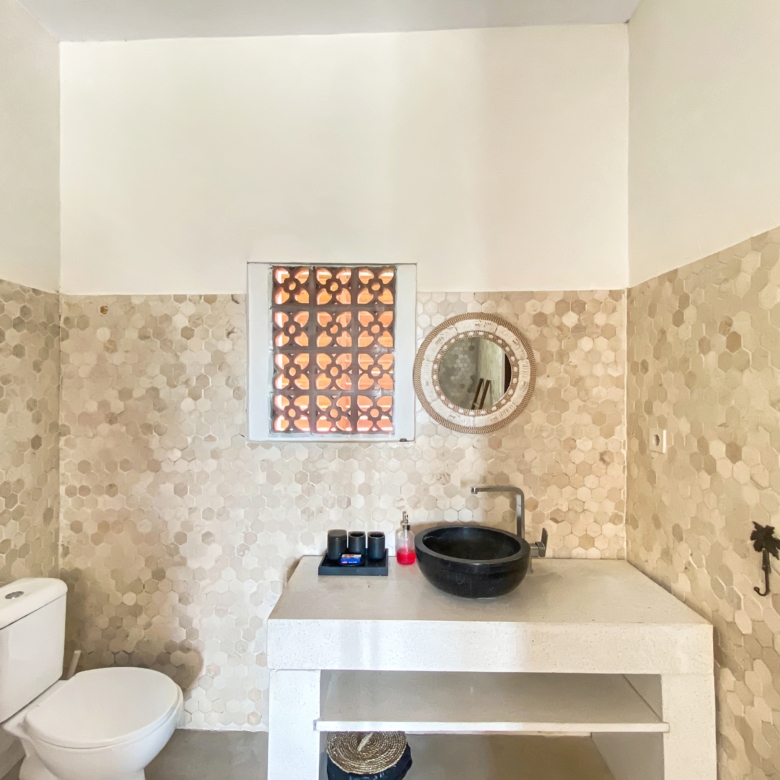 Bathroom with beige hex tile walls, white countertop, and a black vessel sink; round wicker mirror and small lattice window above the sink.