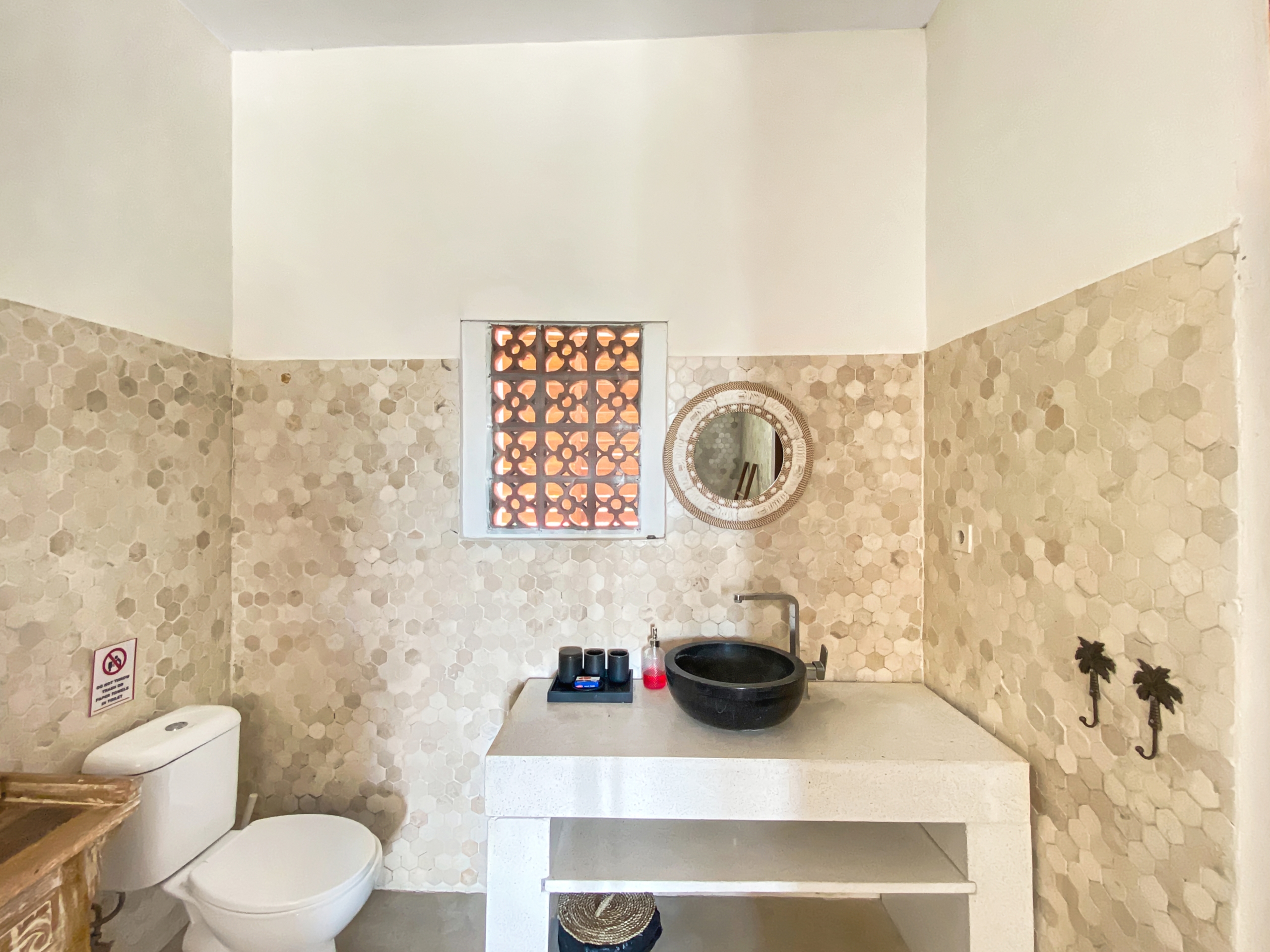 Bathroom with beige hex tile walls, white countertop, and a black vessel sink; round wicker mirror and small lattice window above the sink.