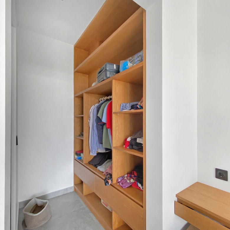 Tall wooden open closet with multiple shelves holding folded clothes and hanging shirts in a bright room