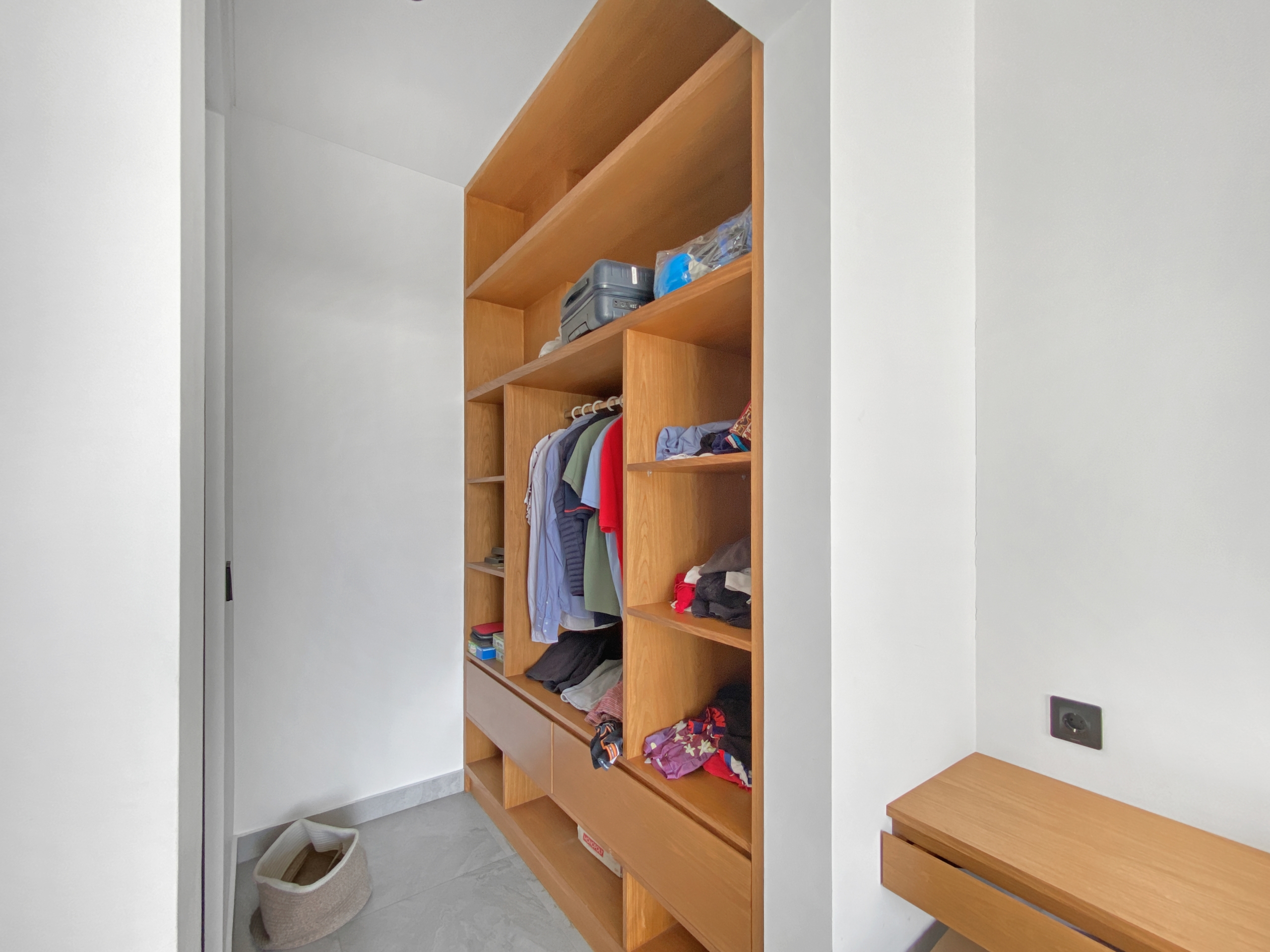Tall wooden open closet with multiple shelves holding folded clothes and hanging shirts in a bright room