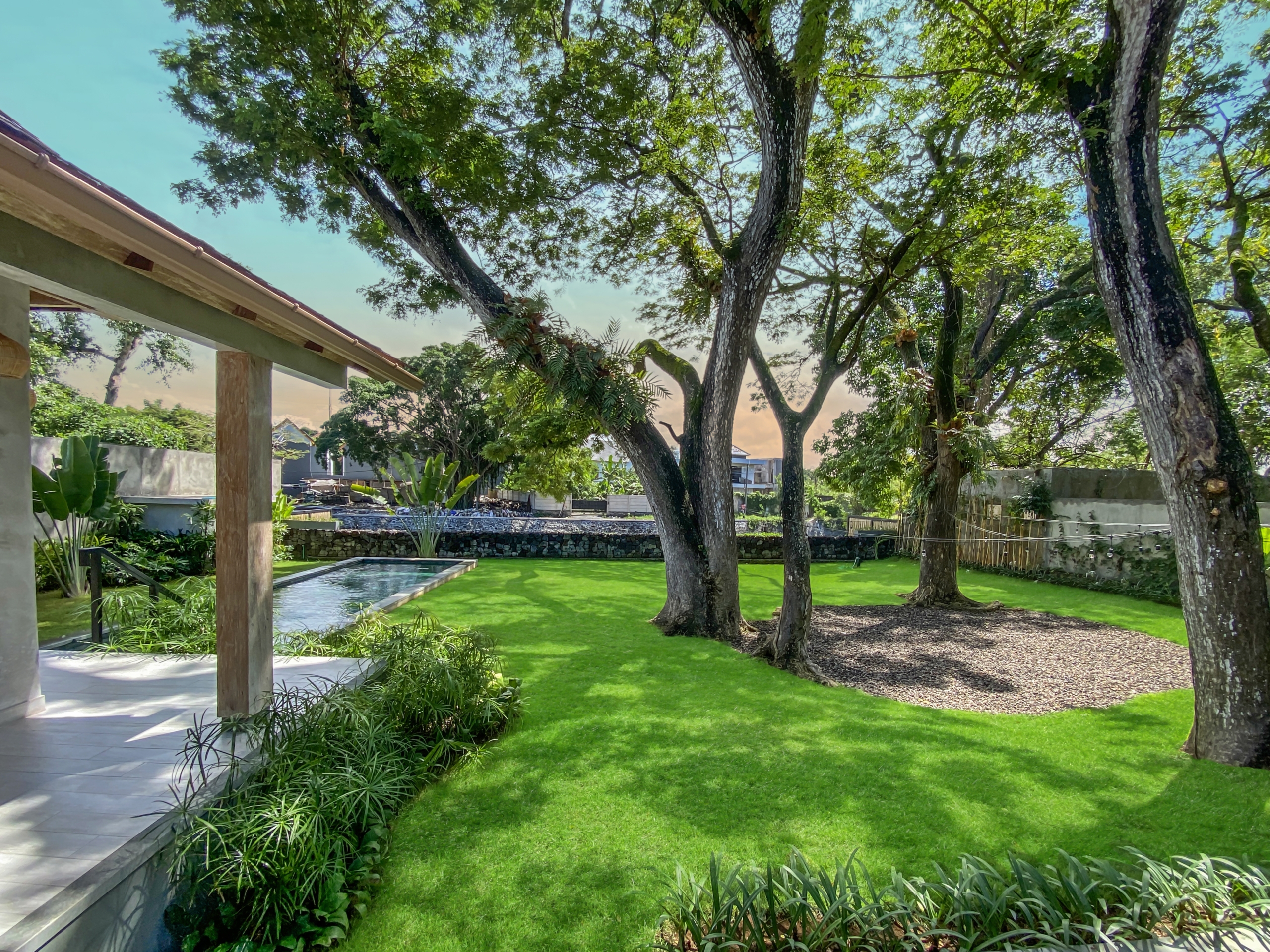 60e7222e16d18ab41fc336c7413e02f249e03ab2 Residential backyard with a green lawn, mature trees, and a rectangular pool beside a stone-and-patio area on the left.