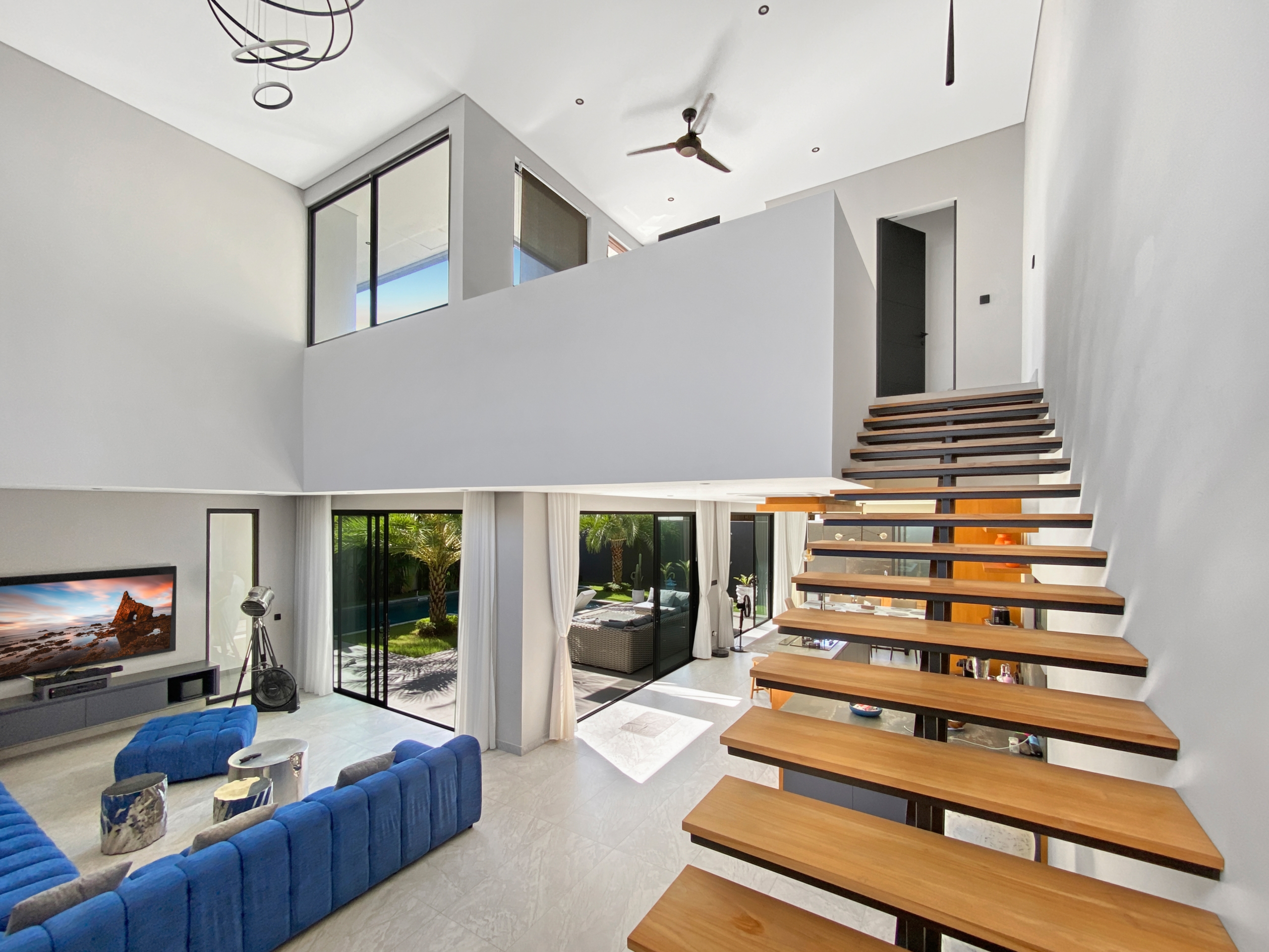 Bright open living room with floating wooden staircase, blue tufted sofa, and glass doors opening to a patio garden.