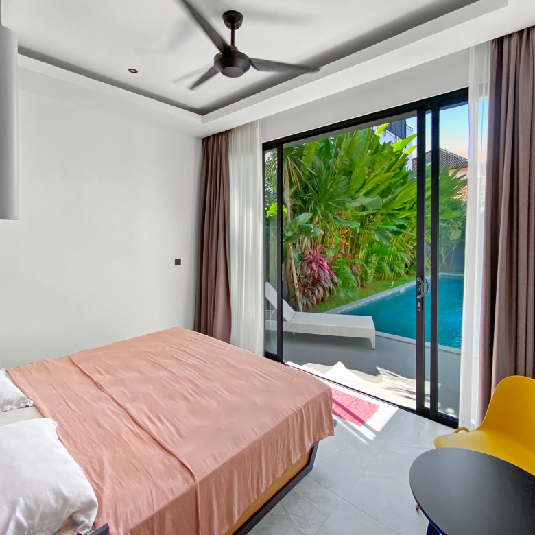 Bedroom with peach bedspread and white pillows, sliding glass doors open to a pool and tropical garden outside.