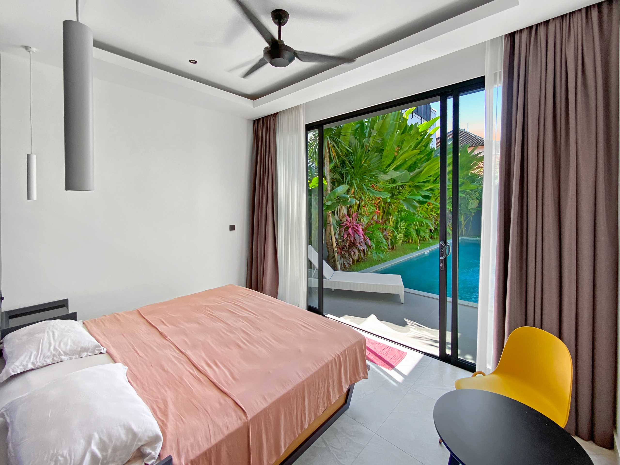 Bedroom with peach bedspread and white pillows, sliding glass doors open to a pool and tropical garden outside.