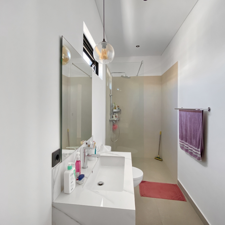 Bright modern bathroom with a wide mirror over a white sink and vanity, shower enclosure in back, and a purple towel hanging on the right.
