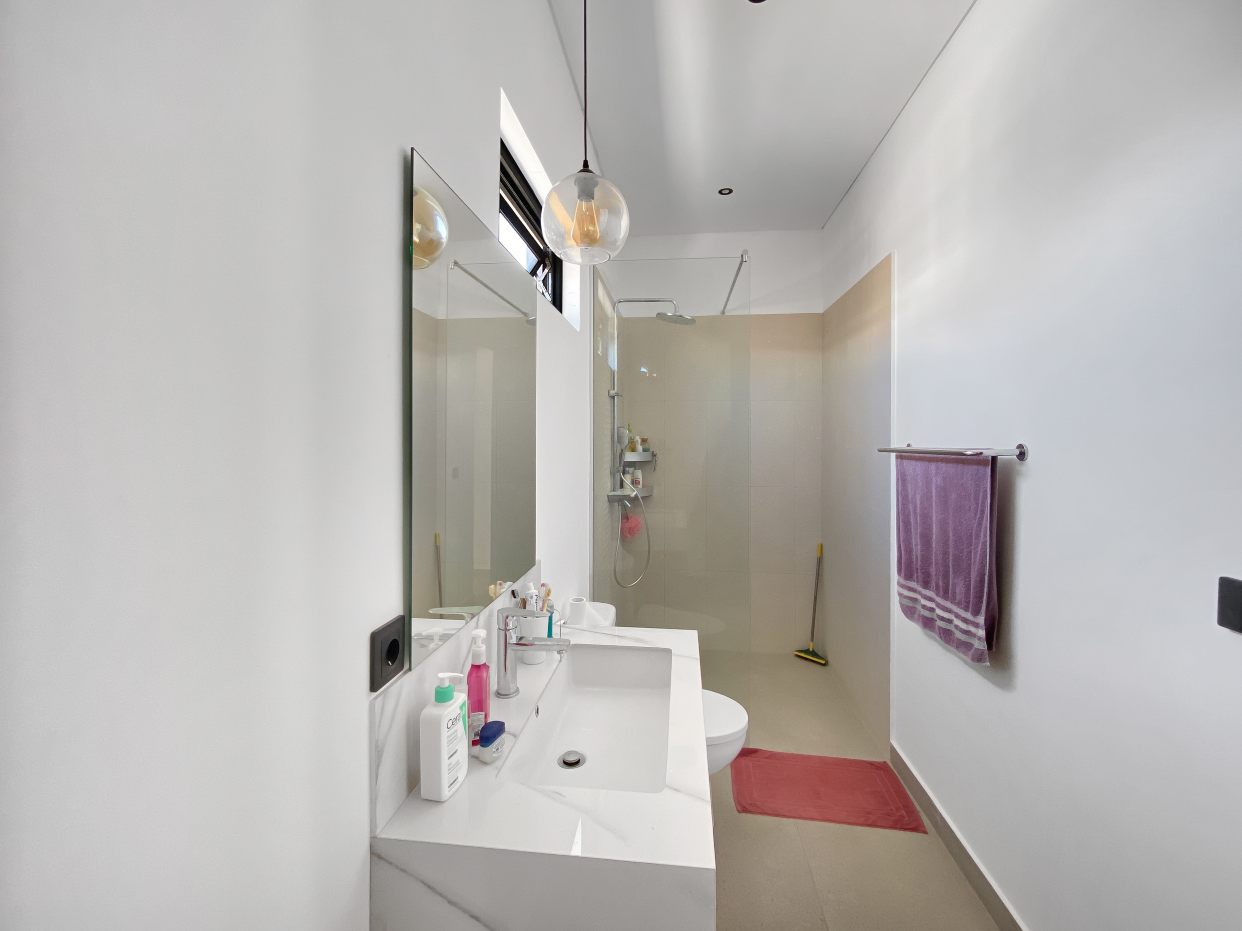 Bright modern bathroom with a wide mirror over a white sink and vanity, shower enclosure in back, and a purple towel hanging on the right.