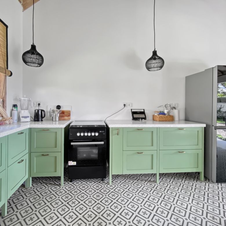 Bright kitchen with mint-green cabinets, a central black oven, two black woven pendant lights, and a patterned tile floor beside a window with a blind on the left.