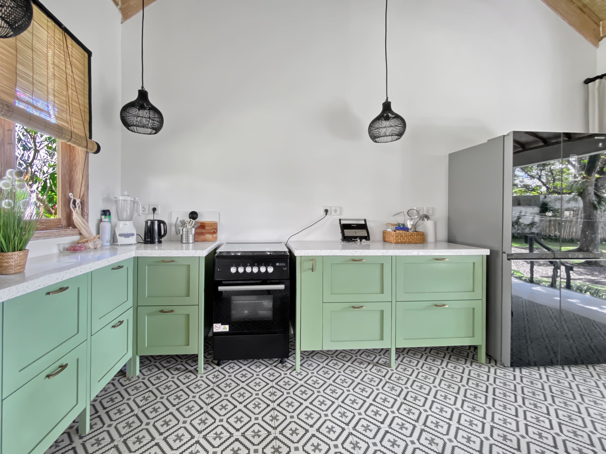6d7efde7c39913a19b8ed7fe287ab210dbabd36a Bright kitchen with mint-green cabinets, a central black oven, two black woven pendant lights, and a patterned tile floor beside a window with a blind on the left.