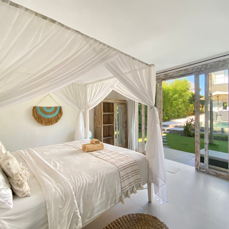 Bright bedroom with two white canopy beds, linen bedding, and towels, opening to a sunny garden through glass doors.