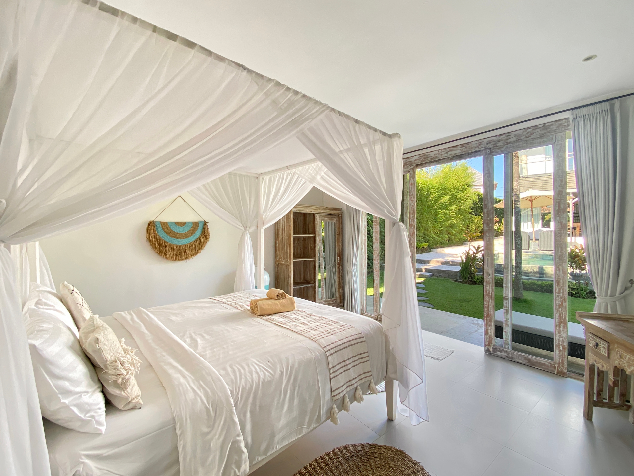 Bright bedroom with two white canopy beds, linen bedding, and towels, opening to a sunny garden through glass doors.