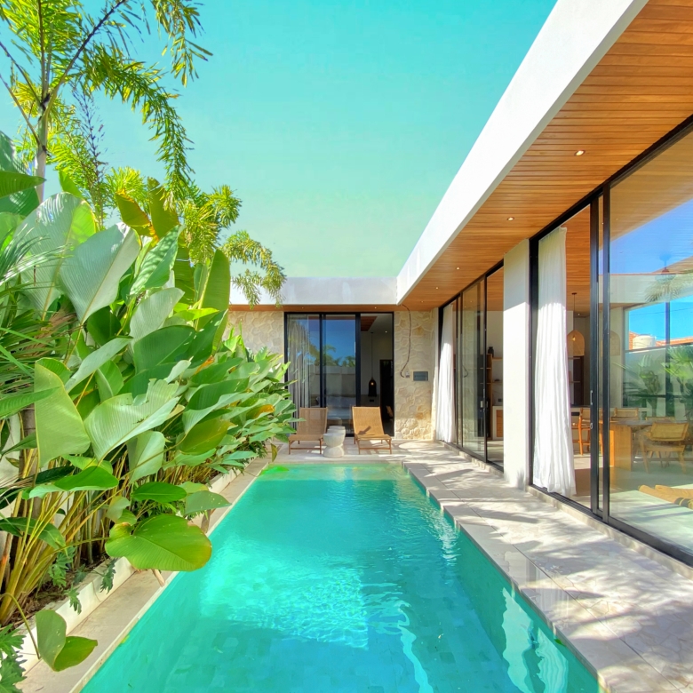 Modern villa poolside: turquoise rectangular pool beside glass-walled living area and a wooden ceiling, with lush tropical plants on the left.