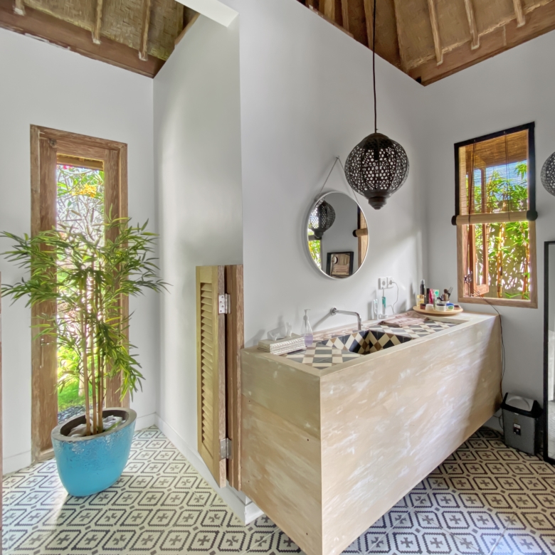 Modern bathroom vanity area with a light wood cabinet, round mirror, and two decorative pendant lights; plant near window adds greenery.