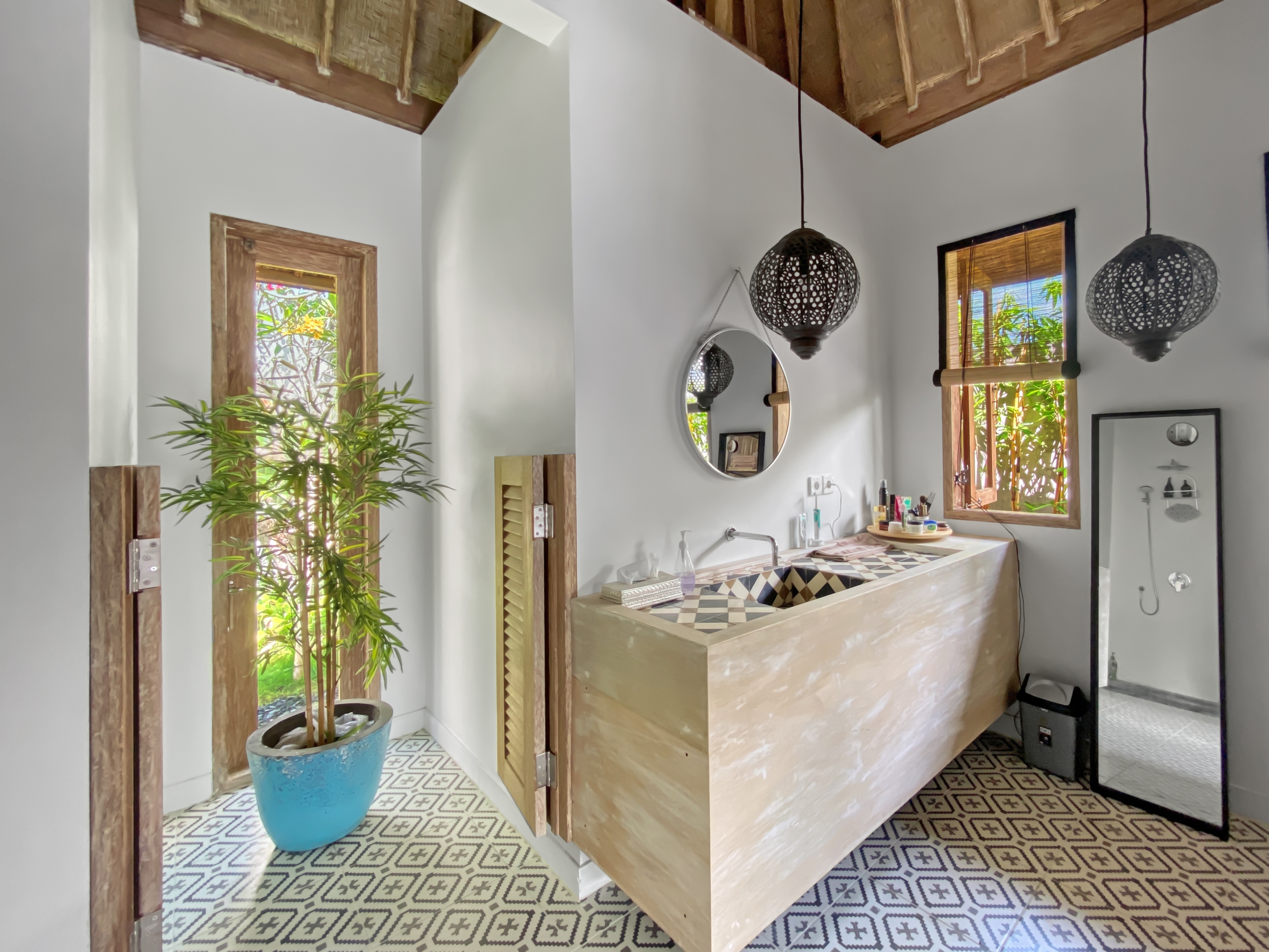 7f40413237f6322ad27a2a40ee6fb552529d1d30 Modern bathroom vanity area with a light wood cabinet, round mirror, and two decorative pendant lights; plant near window adds greenery.