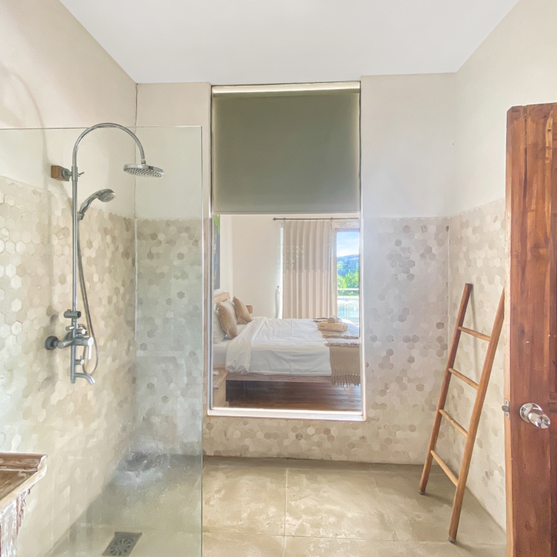Glass shower enclosure in a modern bathroom with hex tile walls and a wooden door on the right; bedroom is visible in the reflection.