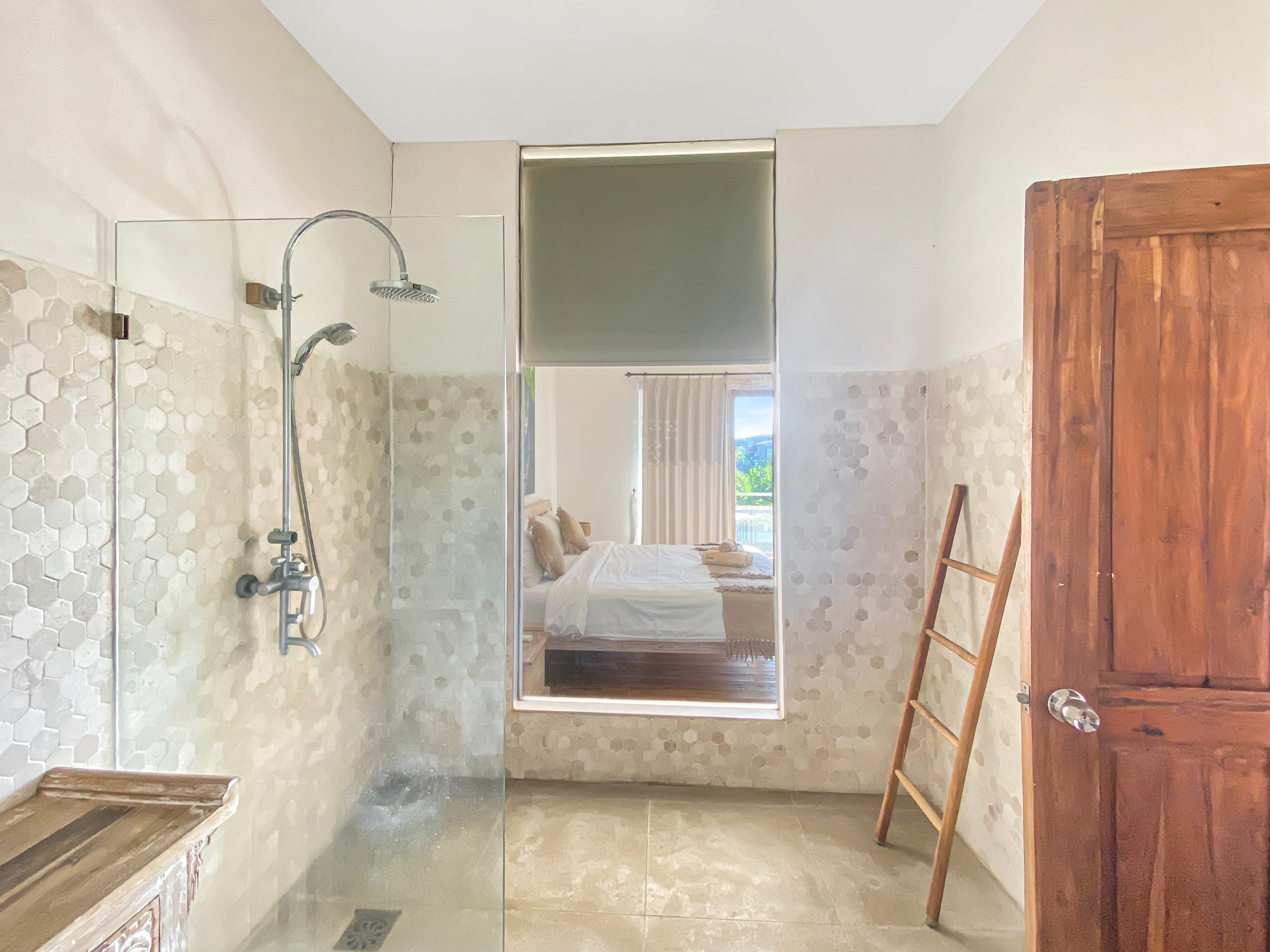 Glass shower enclosure in a modern bathroom with hex tile walls and a wooden door on the right; bedroom is visible in the reflection.