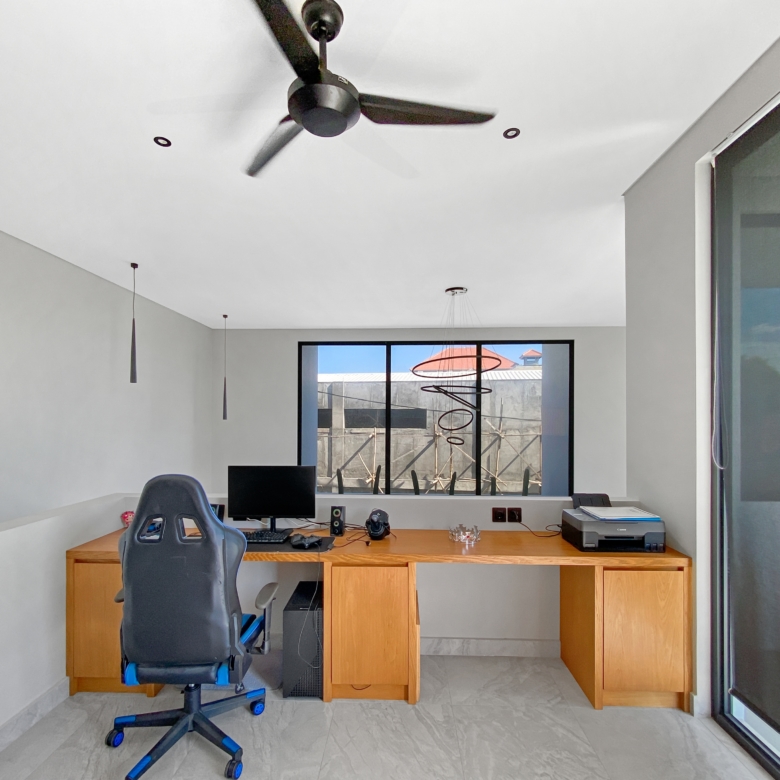 Home office with a wooden desk, computer setup, and a black ergonomic chair; a ceiling fan hangs from the white ceiling above. Clear window at back shows outdoor spiral sculpture.