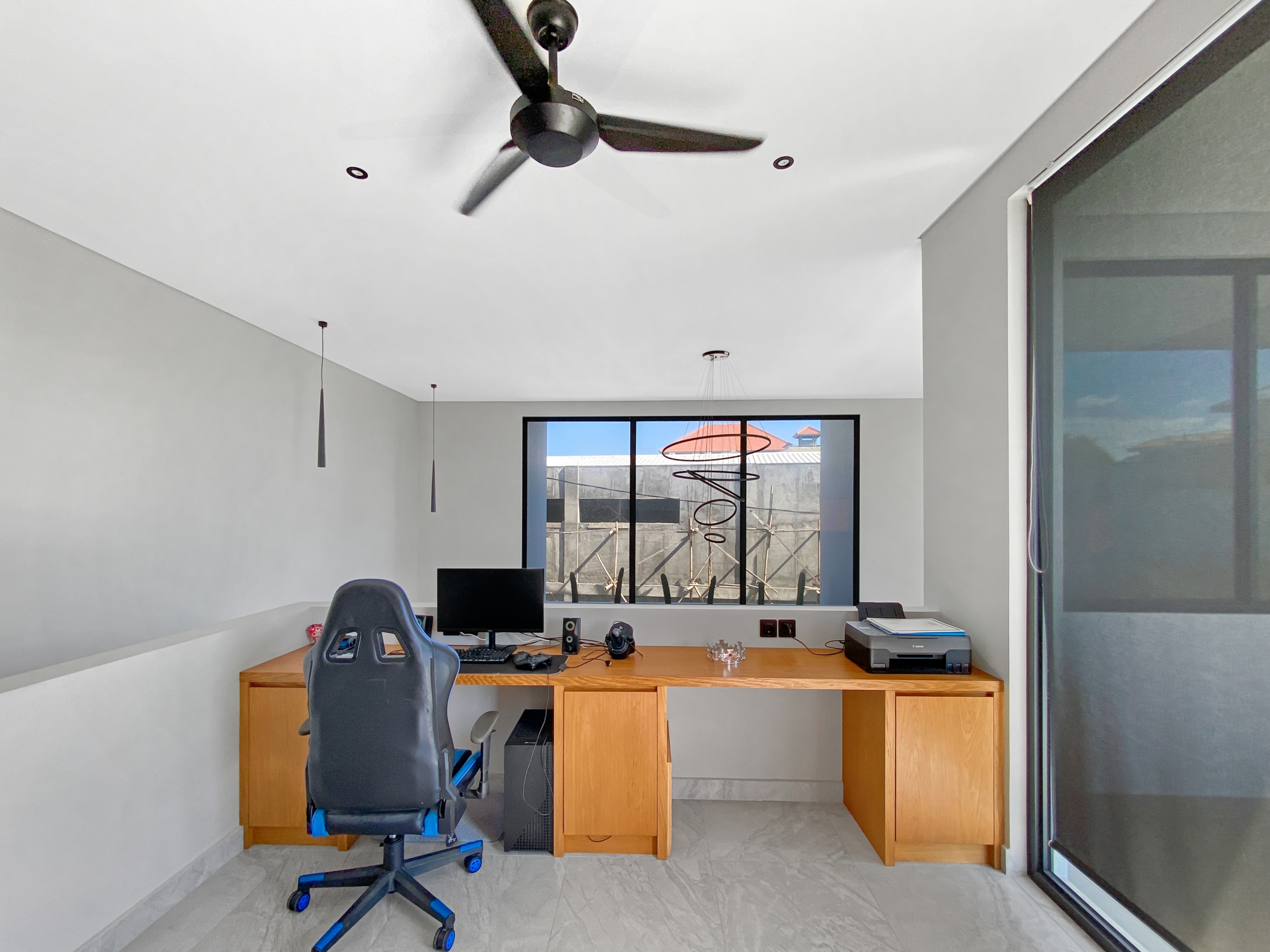 Home office with a wooden desk, computer setup, and a black ergonomic chair; a ceiling fan hangs from the white ceiling above. Clear window at back shows outdoor spiral sculpture.