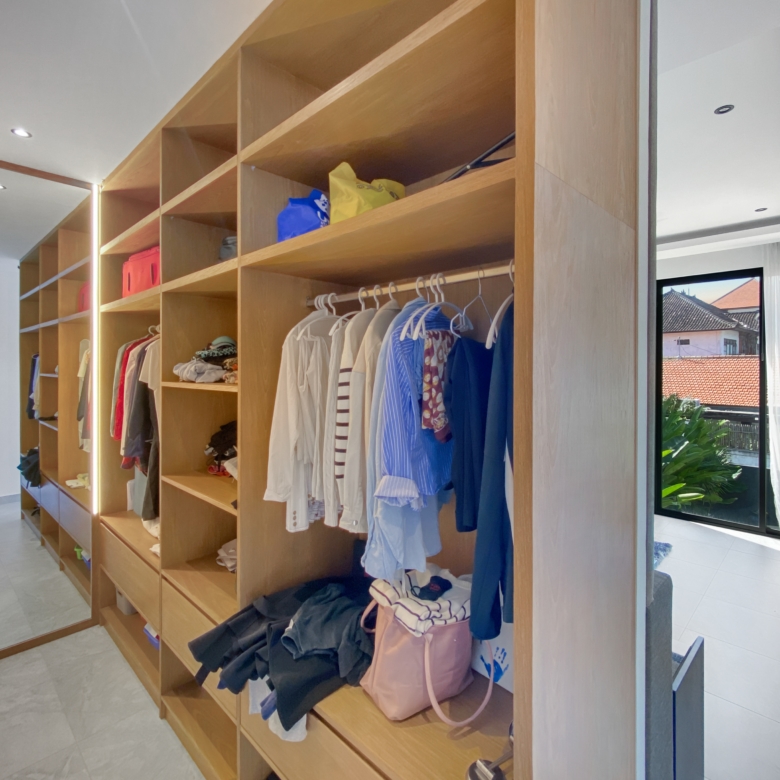 Walk-in closet with tall wooden shelves, clothes hanging on a rail and bags on the shelves in a bright room with a mirror on the left side.