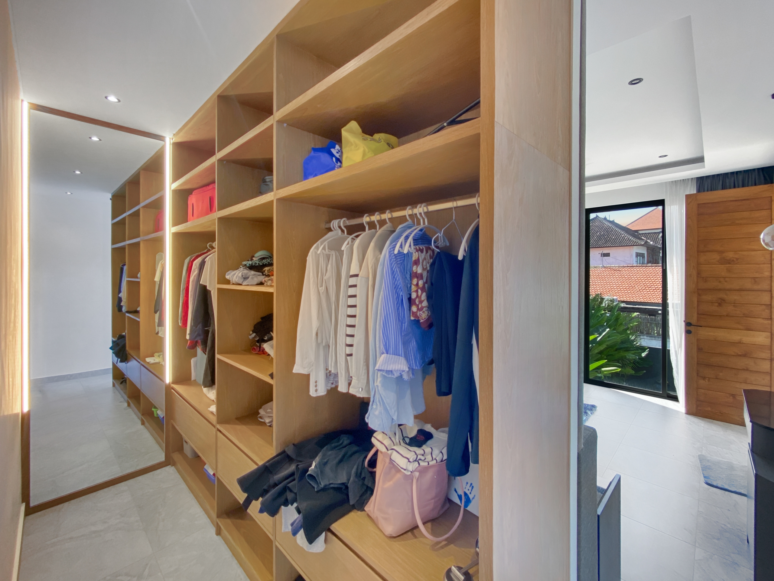 Walk-in closet with tall wooden shelves, clothes hanging on a rail and bags on the shelves in a bright room with a mirror on the left side.