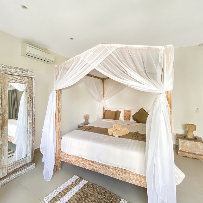 Four-poster bed with white draped canopy and matching linens in a neutral-toned bedroom with rustic wooden furniture and towels arranged on the bed.