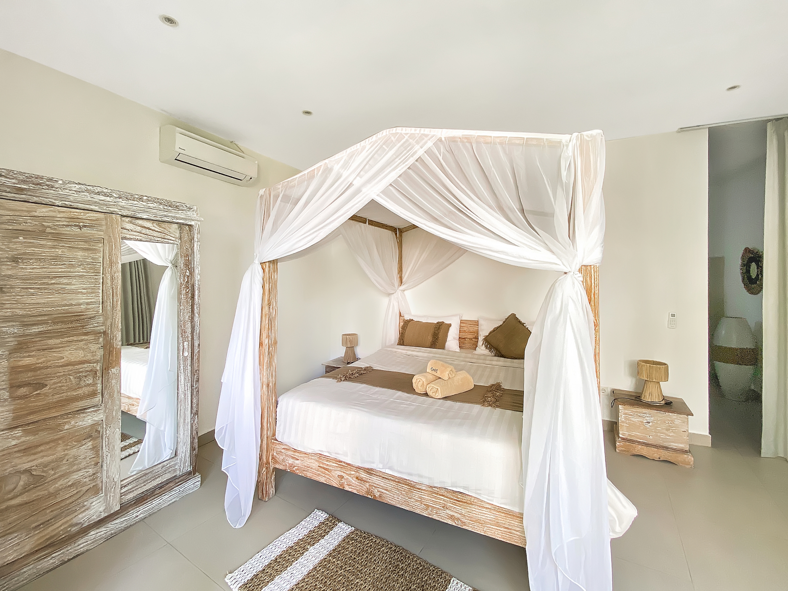 Four-poster bed with white draped canopy and matching linens in a neutral-toned bedroom with rustic wooden furniture and towels arranged on the bed.