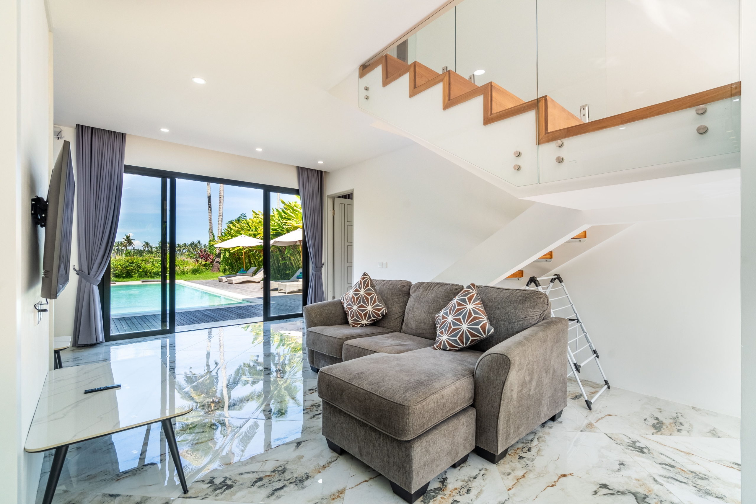 Modern living room with a gray sofa and patterned throw pillows, glass doors to a pool patio outside.
