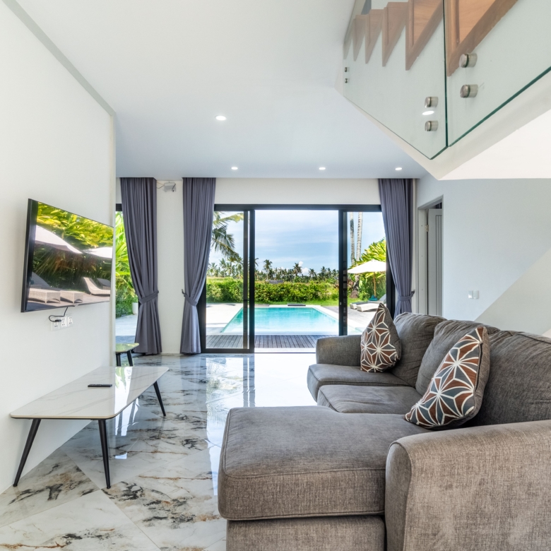 Modern living room with grey sectional, patterned throw pillows, and marble floor; large glass doors open to a sunny pool area outside.