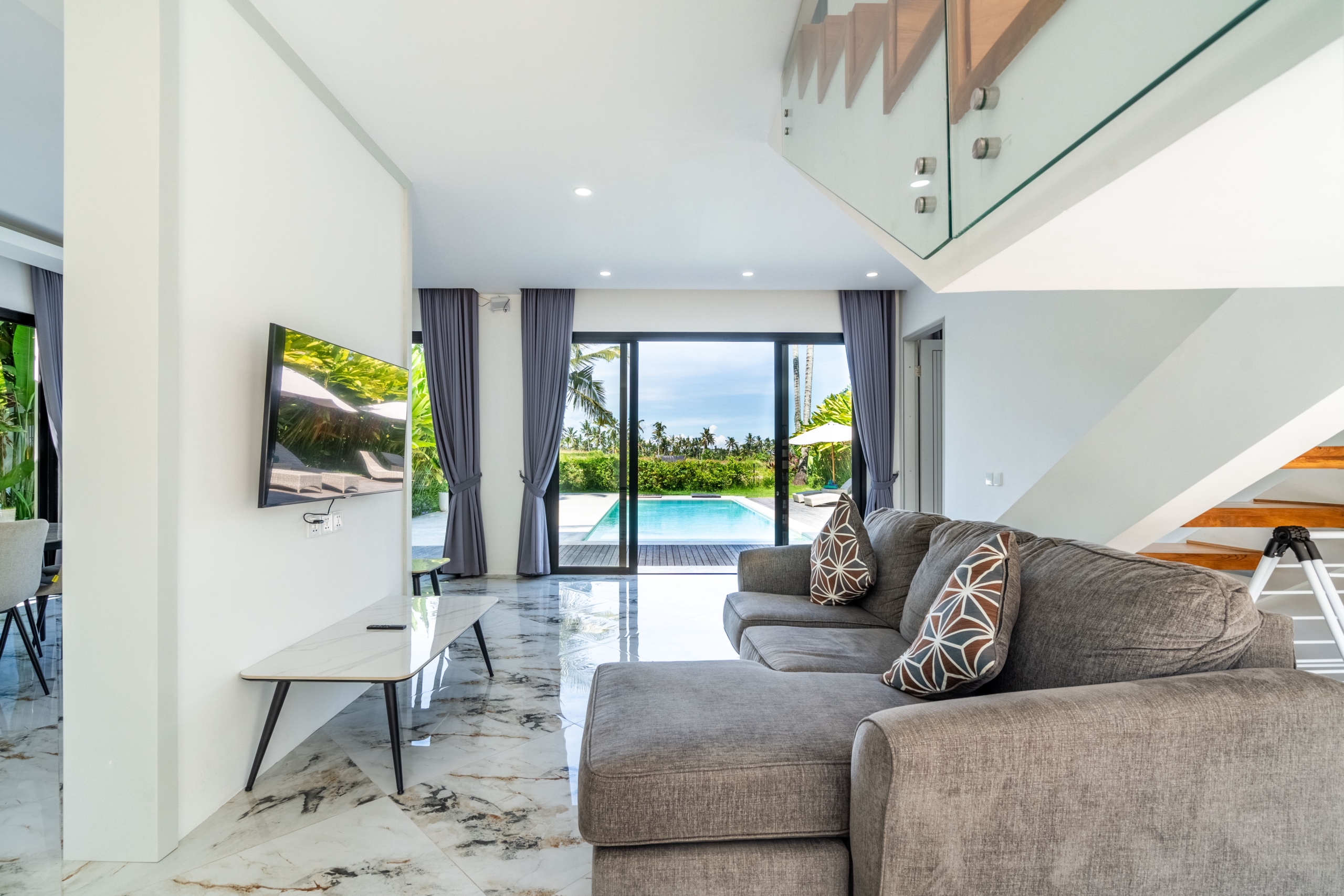 Modern living room with grey sectional, patterned throw pillows, and marble floor; large glass doors open to a sunny pool area outside.