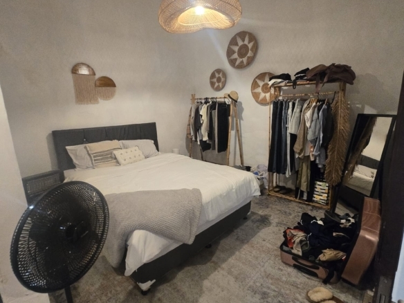 Bedroom with a bed, woven pendant light, and a clothing rack full of hanging clothes; suitcase and slippers on the floor nearby.