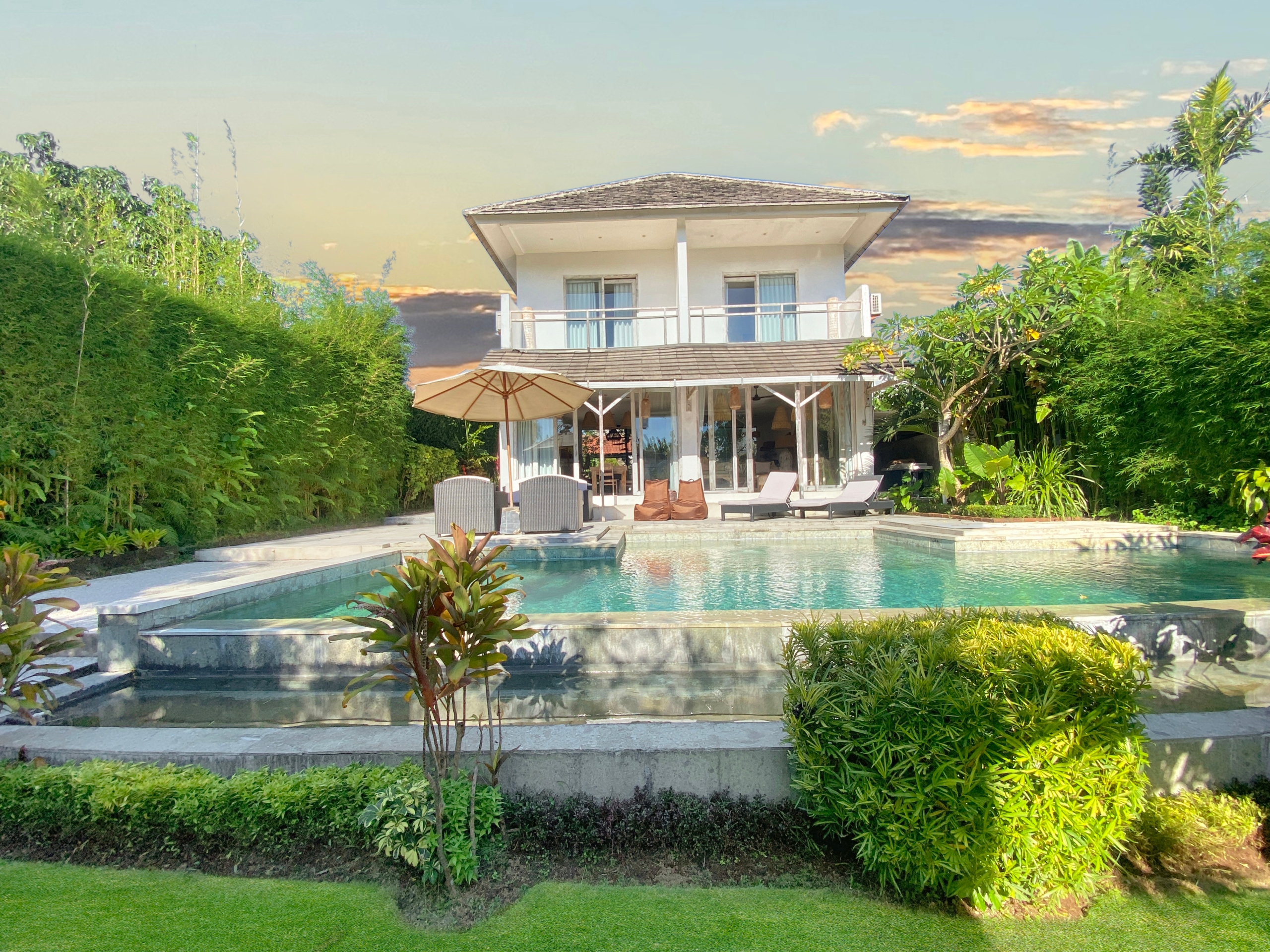 Two-story white villa with a balcony, set behind a rectangular pool and tropical greenery.