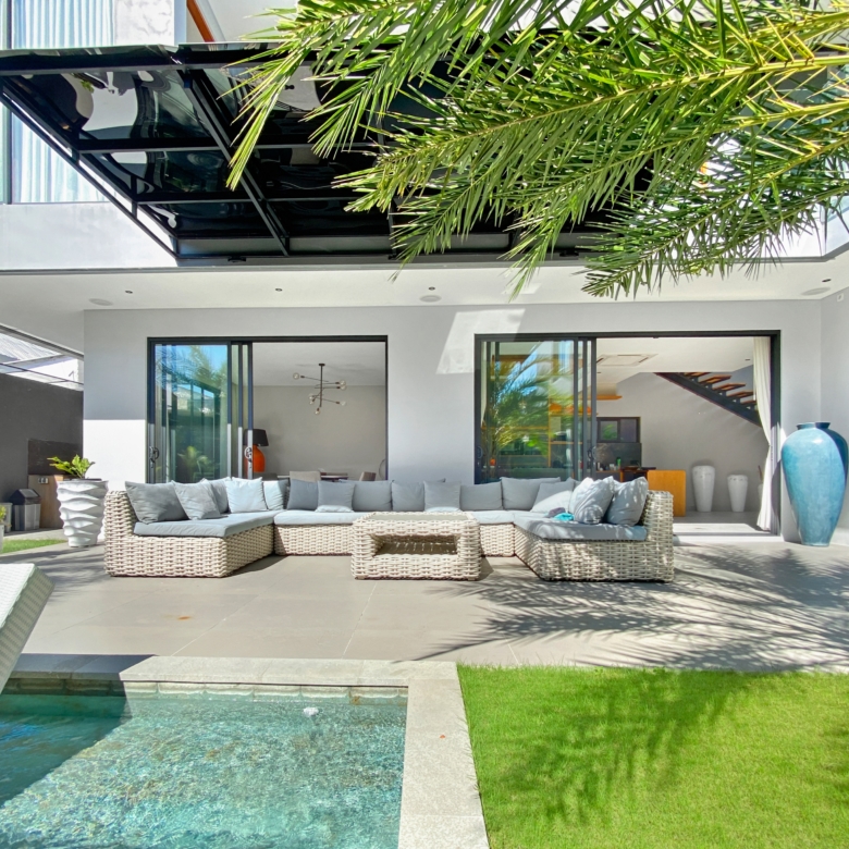 Modern outdoor living area with glass doors, a gray sectional of wicker furniture, and palm fronds overhead.