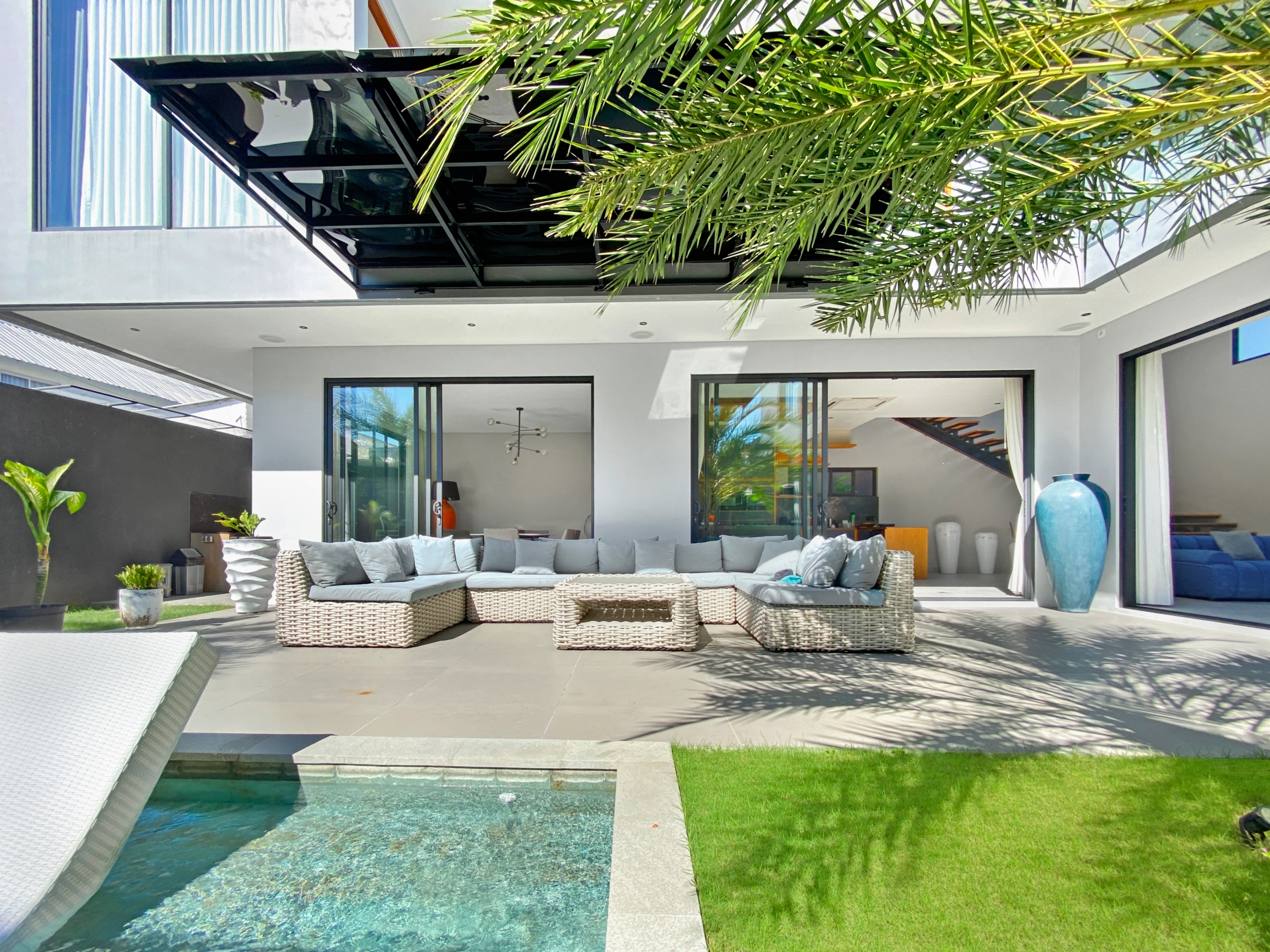 Modern outdoor living area with glass doors, a gray sectional of wicker furniture, and palm fronds overhead.