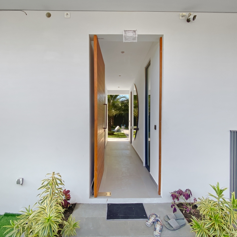 Open doorway in white wall reveals a bright hallway leading to an outdoor courtyard with palm trees; interior shows a wooden door on the left.